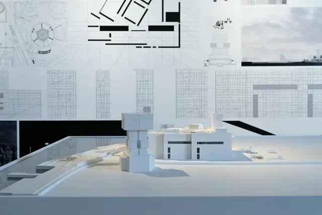 A white architectural massing model on a pale surface, set against technical drawings pinned behind it.